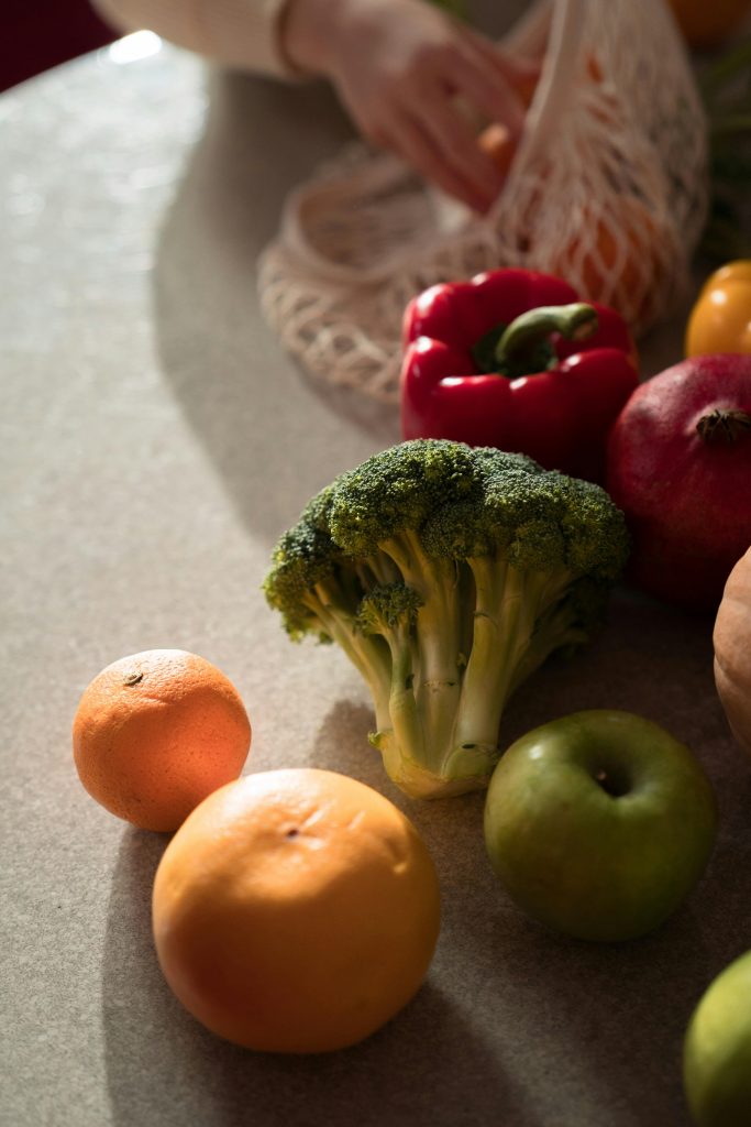Assortment of fresh fruits and vegetables including broccoli, oranges, apple, pepper on kitchen counter.